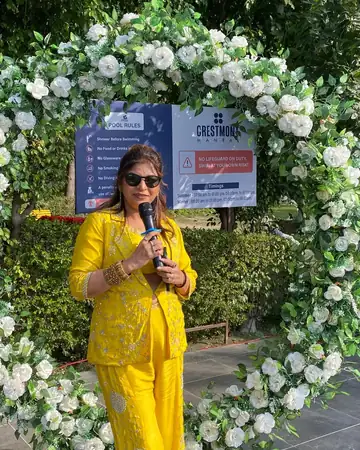 Anu Manocha with mic at a haldi ceremony flower arch