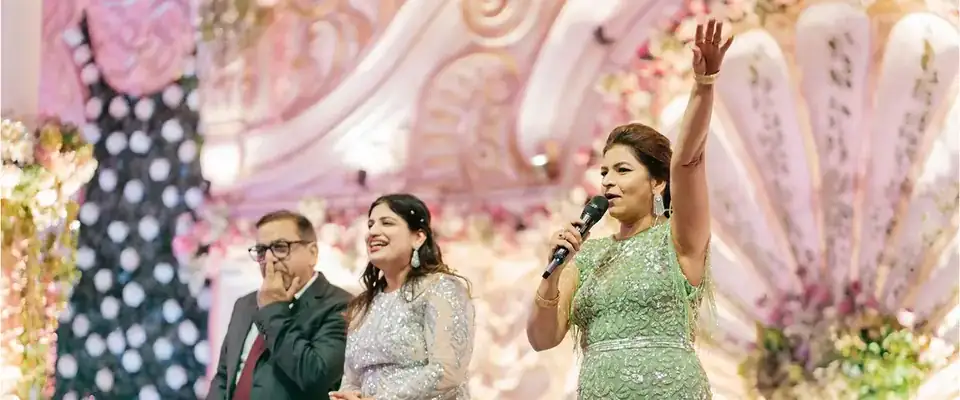 Anchor Anu Manocha on a wedding sangeet stage with the bride's parents, mic in hand and arm raised