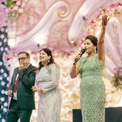 Anchor Anu Manocha on a wedding sangeet stage with the bride's parents, mic in hand and arm raised