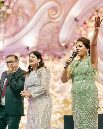 Anchor Anu Manocha on a wedding sangeet stage with the bride's parents, mic in hand and arm raised